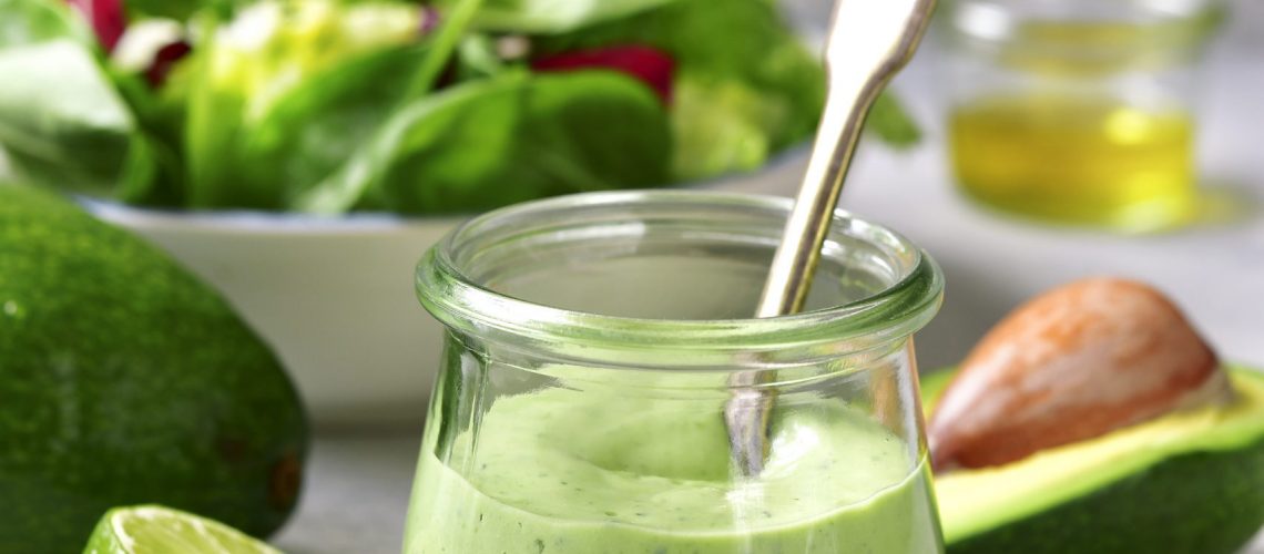 Homemade avocado yogurt dressing in a vintage glass jar with ingredients for making on a light slate, stone or concrete background.